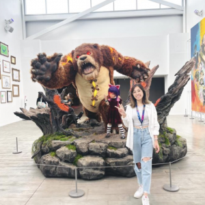 A woman standing in front of a video game statue smiling and holding up two fingers in a peace sign 